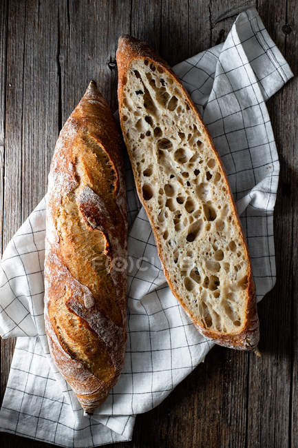 From above whole and halved appetizing baguettes arranged on white towel against rustic wooden background. — Stock Photo