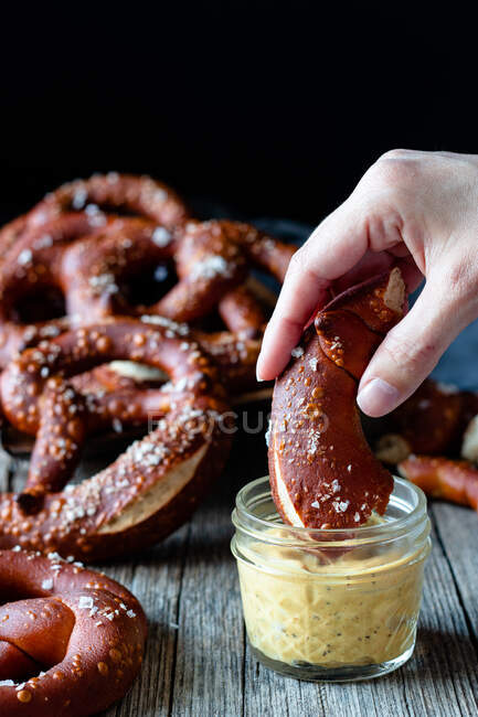 Persona de la cosecha sumergiendo pretzel fresco casero con sal en salsa de queso en la mesa de madera - foto de stock
