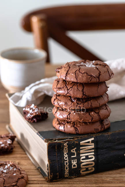 Pilha de biscoitos de chocolate caseiros e livro de receitas em mesa de madeira contra fundo turvo — Fotografia de Stock