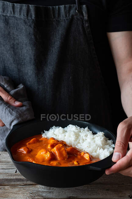 Persona de la cosecha en delantal de pie junto a la mesa de madera con sartén llena de deliciosa mantequilla de pollo y arroz - foto de stock
