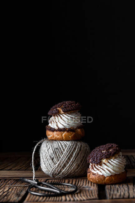 Homemade choux pastries with cream and chocolate scissors and ball of yarn arranged on texture wooden surface against black background — Stock Photo