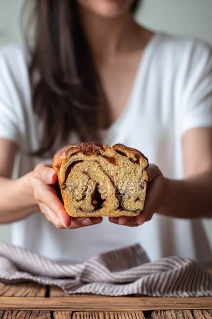 Crop female cook holding fresh twisted bread or cinnamon babka over wooden table with striped towel on blurred background — Stock Photo