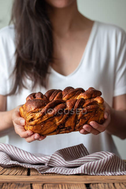 Crop cocinera femenina sosteniendo pan fresco retorcido o babka canela sobre mesa de madera con toalla rayada sobre fondo borroso - foto de stock