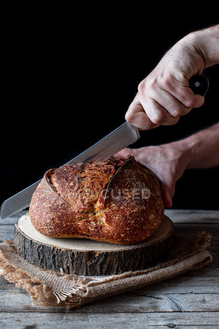 Unbekannter schneidet mit Messer Laib frisches Sauerteigbrot auf Stück Holz vor schwarzem Hintergrund — Stockfoto