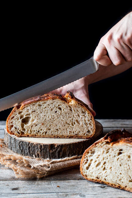 Неупізнавана людина використовує ніж, щоб зрізати буханець свіжого хліба на дереві на чорному фоні. — стокове фото