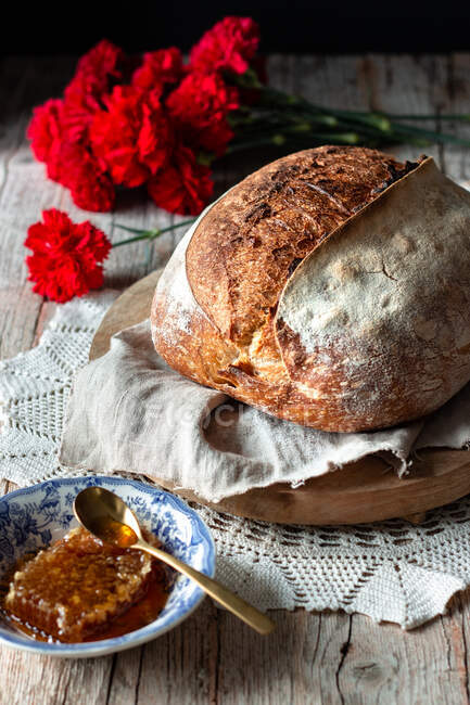 Pane fresco di pasta madre messo a bordo e tovaglioli vicino a nido d'ape e mazzo di garofani rossi su tavola rustica — Foto stock