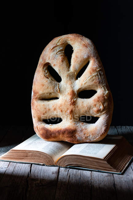 Loaf of fresh fougasse bread placed on open recipe book on wooden table against black background — Stock Photo