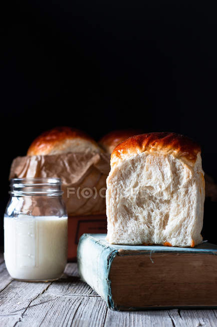 Small jar of fresh milk and buns of baked bread on old recipe book placed on timber table. — Stock Photo