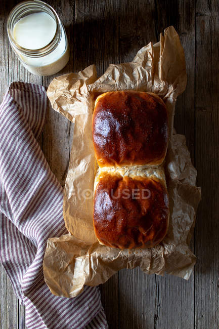 Dall'alto panini di pane fresco su pergamena messi vicino a vaso di latte fresco e tovagliolo su tavolo di legname . — Foto stock