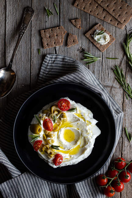 Top vie wof piatto di yogurt labneo con pomodori e olive su tavolo di legno vicino tovagliolo, cucchiaio e cracker croccanti e rosmarino — Foto stock