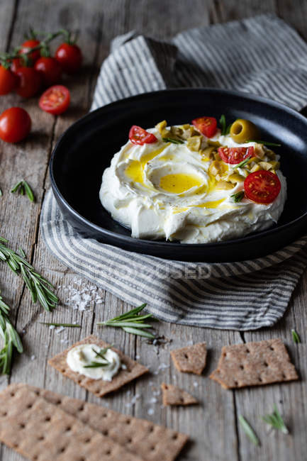 Ciotola di yogurt labneo con pomodori e olive su tavola di legno vicino tovagliolo e cracker croccanti e rosmarino — Foto stock