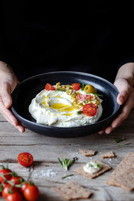 Ciotola di yogurt labneo con pomodori e olive in mani di chef maschio su tavola di legno — Foto stock