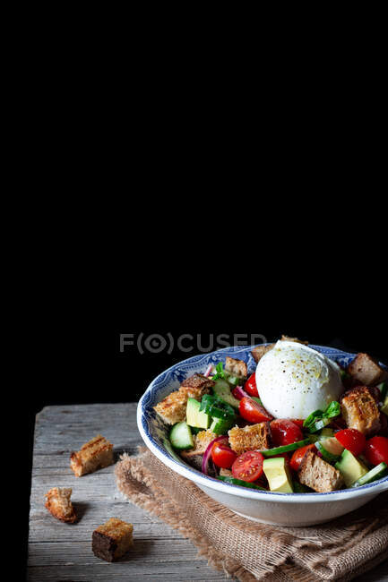 Bowl with yummy panzanella salad placed on cloth on wooden table against black background — Stock Photo