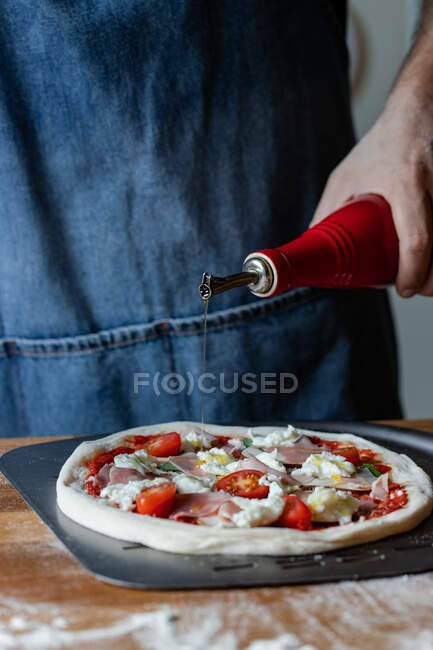 Невпізнаваний чоловік в фартусі розливає оливкову олію зверху піци, стоячи за столом і готуючи на кухні — стокове фото