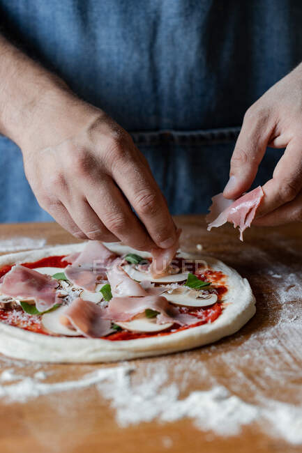 Unrecognizable chef in apron putting tasty ham on raw pizza while cooking on table in kitchen — Stock Photo