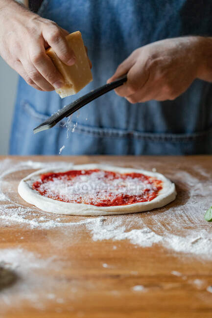 Невпізнаваний хлопець в фартусі шліфує свіжий сир на тісті з томатним соусом, готуючи піцу на столі — стокове фото