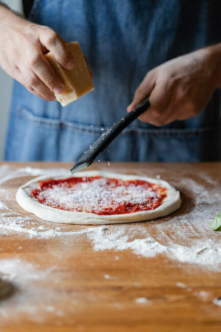 Unbekannter in Schürze zermahlt Frischkäse auf Teig mit Tomatensauce bei der Zubereitung von Pizza auf dem Tisch — Stockfoto