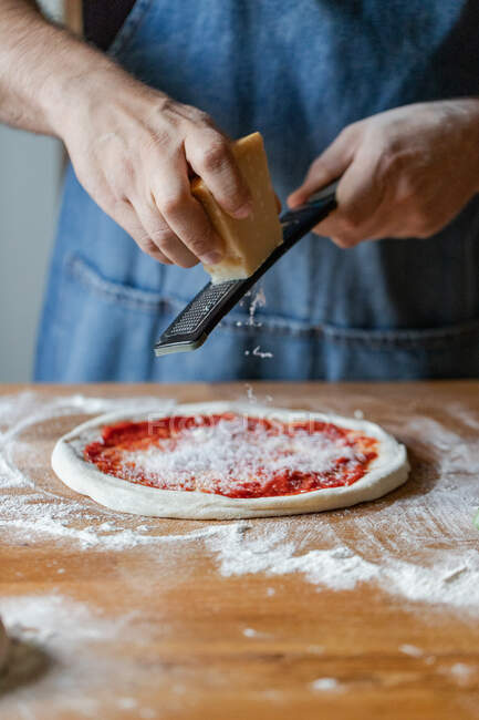 Неузнаваемый парень в фартуке, шлифующий свежий сыр на тесте с томатным соусом во время приготовления пиццы на столе — стоковое фото