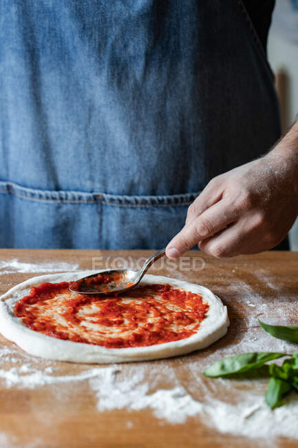 Chef anônimo esfregando molho de tomate fresco na massa crua enquanto cozinha pizza na mesa — Fotografia de Stock