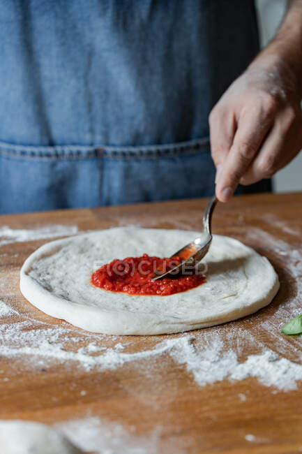 Anonymer Koch schmiert frische Tomatensauce auf rohen Teig, während er Pizza auf dem Tisch kocht — Stockfoto