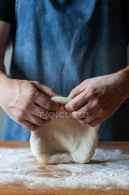 Masculino irreconocible en delantal aplanando masa blanda sobre la mesa con harina mientras cocina pizza - foto de stock