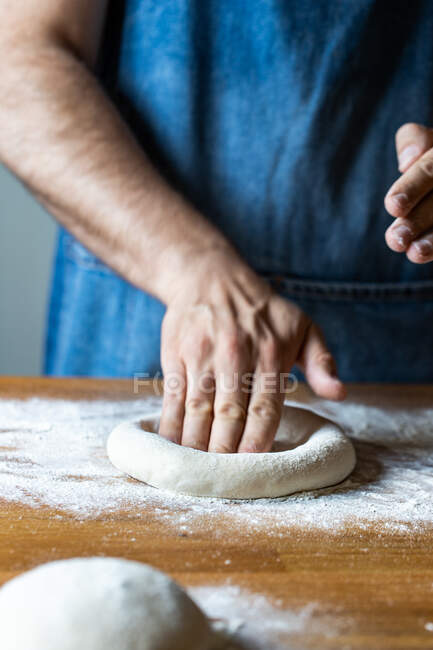 Неузнаваемый мужчина в фартуке сглаживает мягкое тесто за столом с мукой во время приготовления пиццы — стоковое фото