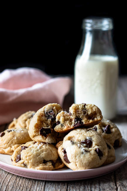 Piatto di gustosi biscotti con gocce di cioccolato poste sul tavolo di legname vicino al latte sfocato e stoffa su sfondo nero — Foto stock