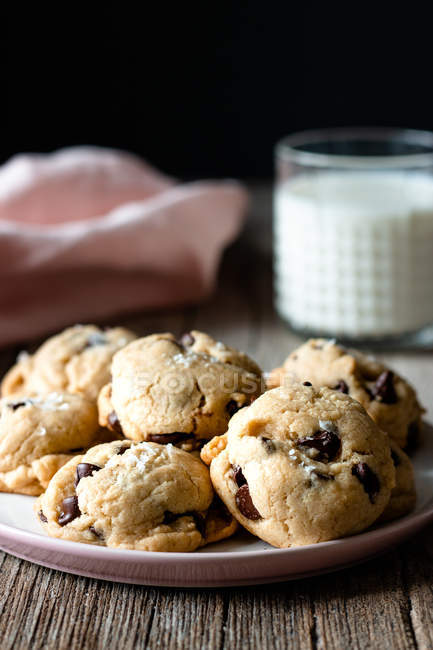 Plate of tasty cookies with chocolate chips placed on lumber table near blurred milk and cloth against black background — Stock Photo