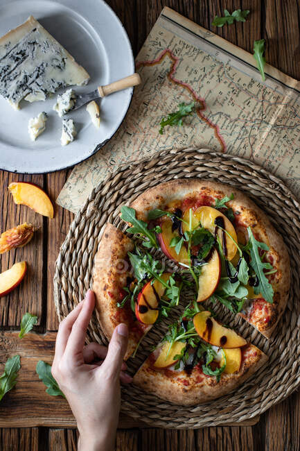 From above unrecognizable person taking piece of yummy summer pizza with peach and arugula near map and cheese on wooden table — Stock Photo