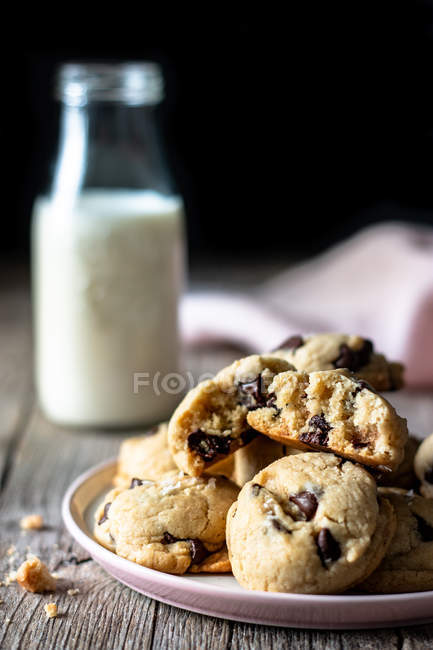 Assiette de biscuits savoureux avec des pépites de chocolat placées sur la table de bois près du lait flou et du tissu sur fond noir — Photo de stock