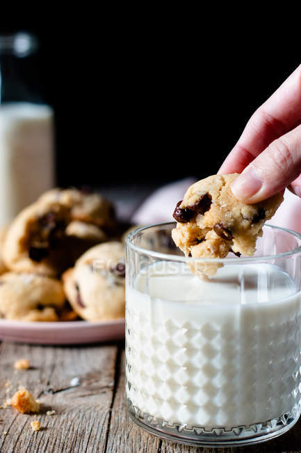Unrecognizable female dipping piece of yummy cookie with chocolate chips into glass of fresh milk on wooden table — Stock Photo