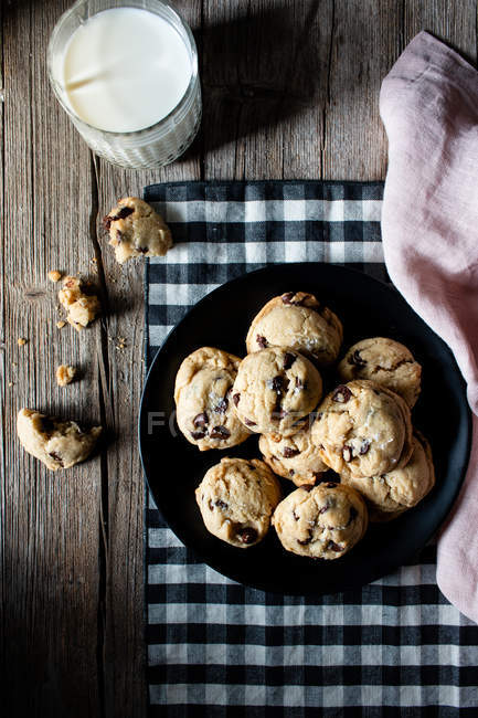 Assiette de biscuits savoureux avec des pépites de chocolat placées sur la table de bois près du lait et du tissu sur la table en bois — Photo de stock