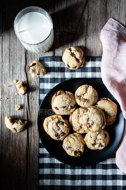 Piatto di gustosi biscotti vegani con gocce di cioccolato poste sul tavolo di legname vicino al latte e al panno sfocati sullo sfondo nero — Foto stock