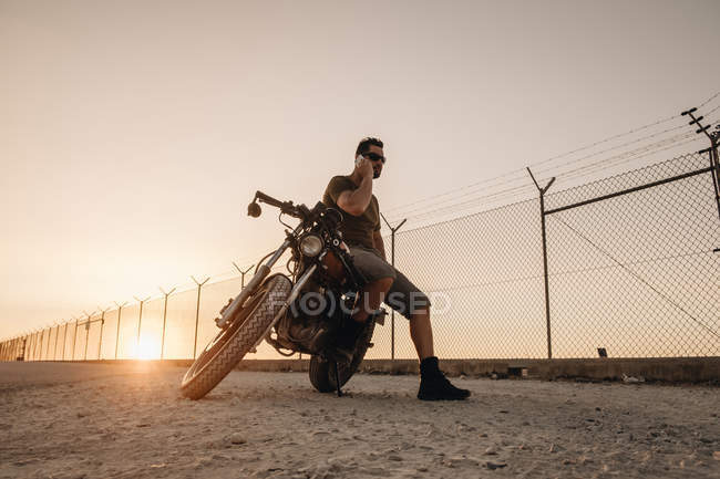 Uomo seduto sulla moto e parlare sul cellulare al tramonto nel campo — Foto stock