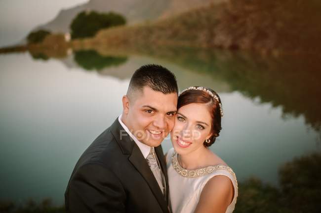 Очаровательная улыбающаяся невеста и жених обнимаются и смотрят в камеру на фоне спокойного хрустального озера — стоковое фото