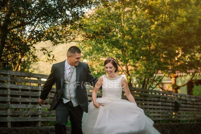 Элегантный веселый жених держит очаровательную невесту и бегает вместе со смехом по дороге вдоль деревянного забора в зеленом саду — стоковое фото