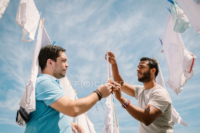 Side view of handsome bearded multi ethnic men hanging white clothes outside on background of blue sky — Stock Photo