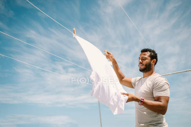 Vista lateral do homem hispânico concentrado fixando t-shirt branca com rotação estreita na corda no fundo do céu brilhante — Fotografia de Stock