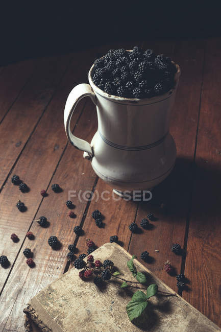 Vintage jag with ripe blackberries on wooden table and leaves on old book — Stock Photo