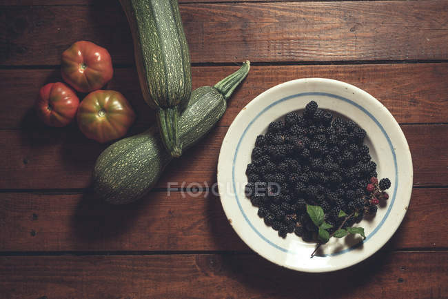 Ripe blackberries garnished by vegetables in bowl on wooden background — Stock Photo