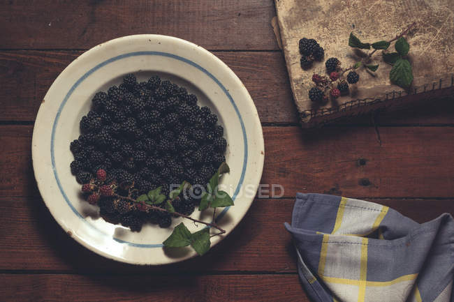 Ripe blackberries garnished by leaves in bowl with vintage book and napkin on wooden table — Stock Photo