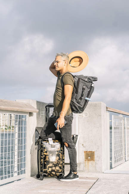 Retrato completo de turista tirando seu chapéu, com mala e mochila em pé na frente do rio — Fotografia de Stock