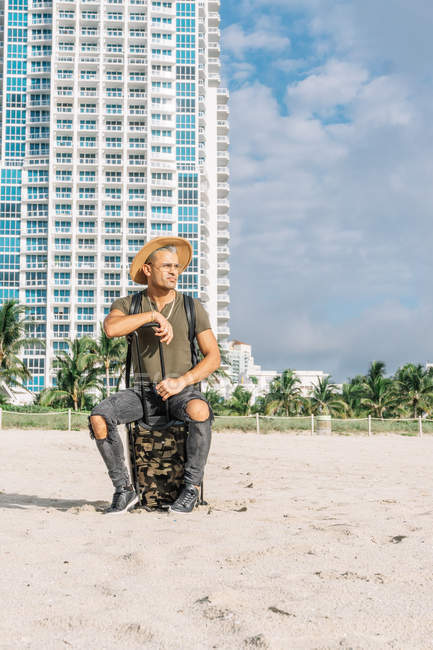Homem atraente sentado em sua bagagem de transporte olhando para longe na praia sul em Miami — Fotografia de Stock