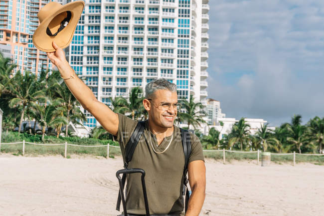 Happy male tourist in hipster clothes waving his hat to someone on south beach — Stock Photo