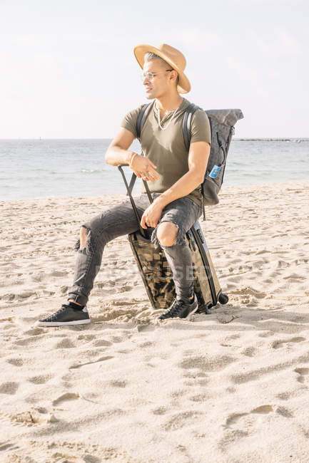 Bonito e ajuste cara posando com pequeno mala no praia contemplando oceano — Fotografia de Stock