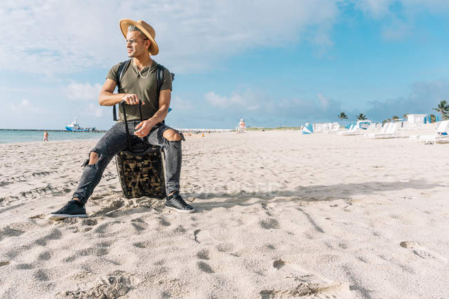 Bonito e ajuste cara posando com pequeno mala no praia contemplando oceano — Fotografia de Stock
