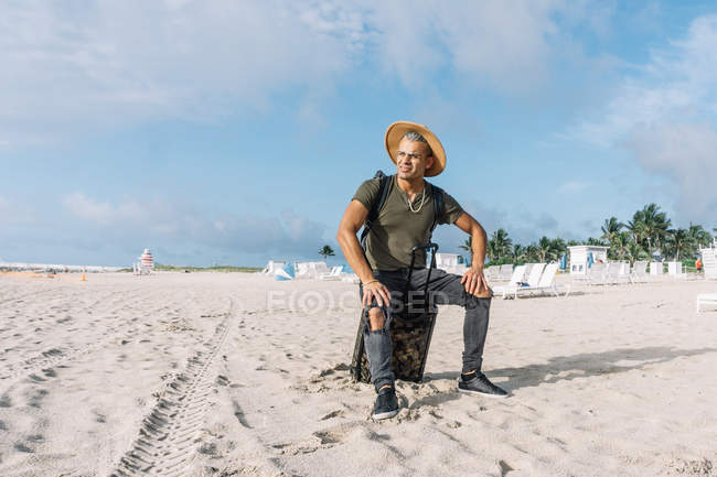 Guapo hipster hombre sentado en el equipaje de mano, hermosa playa, disfrutando de unas vistas impresionantes. - foto de stock