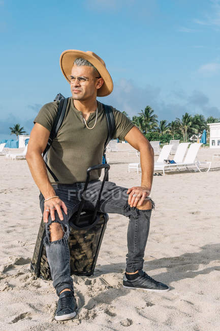 Handsome hipster man sitting on carrying baggage, beautiful beach, enjoying amazing view — Stock Photo