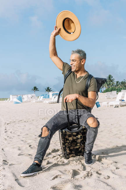Ritratto di allegro giovane che ondeggia ciao seduto sui suoi bagagli nella spiaggia del sud, Miami — Foto stock