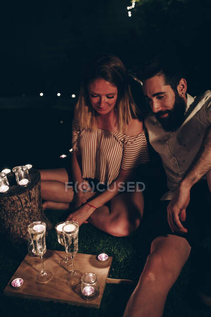 Молодой человек и женщина сидят на лужайке возле светящихся свечей ночью в темном ночном саду — стоковое фото